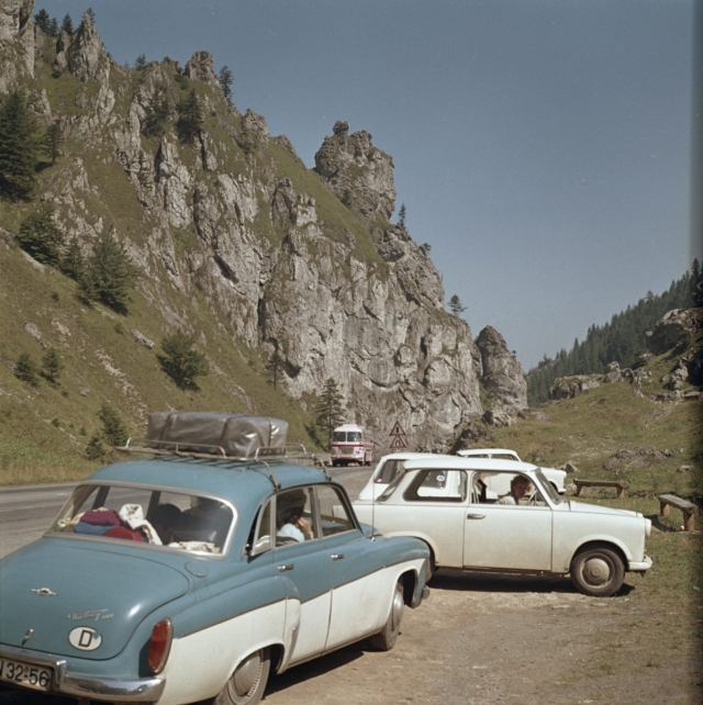 Das Foto zeigt eine typische Urlaubsszene aus der sozialistischen Tschechoslowakei der 1970er Jahre. Im Vordergrund sind zwei charakteristische Autos der Zeit zu sehen - ein türkis-weißer Wartburg (mit Dachgepäckträger) und ein weißer Trabant. Im Hintergrund erkennt man die imposanten Felswände der Kleinen Fatra (Malá Fatra), ein beliebtes Wandergebiet in der heutigen Slowakei. Ein roter Bus ist ebenfalls auf der Bergstraße zu sehen. Das Bild vermittelt einen authentischen Einblick in den Tourismus im damaligen Ostblock, als viele DDR-Bürger ihre Urlaube in den "Bruderländern" verbrachten. Die Aufnahme stammt vom Fotografen Werner Großmann und wurde 1974 gemacht. Die farbigen Felsformationen und die grüne Vegetation der Berglandschaft bilden einen malerischen Hintergrund für diese Momentaufnahme des Urlaubsverkehrs.