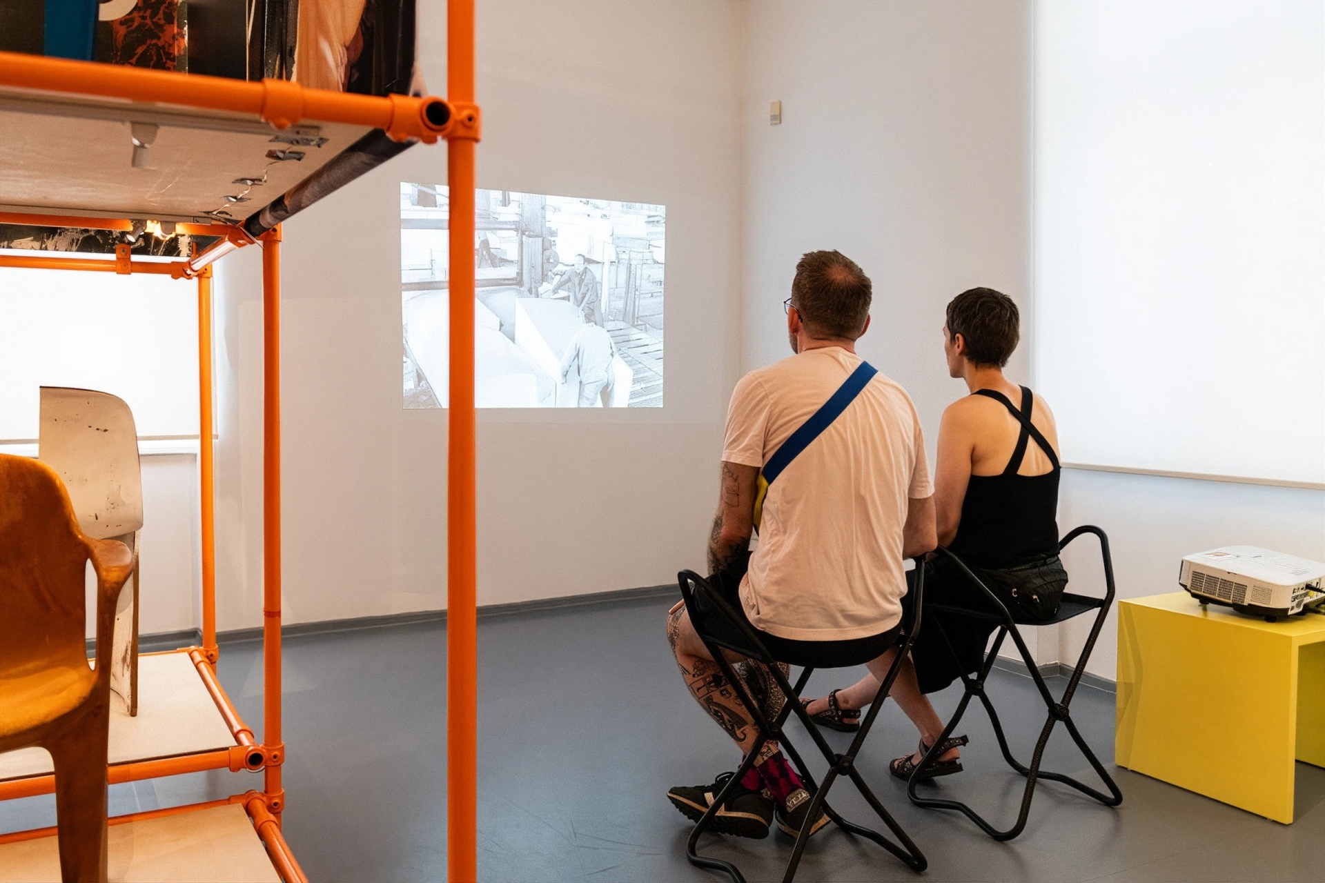 Ausstellungsansicht eines Museumsraums mit zwei Besucher*innen, die auf Klappst&uuml;hlen sitzend eine historische Schwarz-Wei&szlig;-Videoprojektion an der wei&szlig;en Wand betrachten. Im Vordergrund ist ein orangefarbenes Ausstellungsregal zu sehen, auf dem ein Designerstuhl ausgestellt ist. Rechts im Bild steht ein gelber W&uuml;rfel mit einem Projektor. Der Raum ist minimalistisch gestaltet mit grauen Bodenfliesen und wei&szlig;en W&auml;nden. Die Installation vermittelt den dokumentarischen Charakter der Ausstellung zur Design- und Produktionsgeschichte.