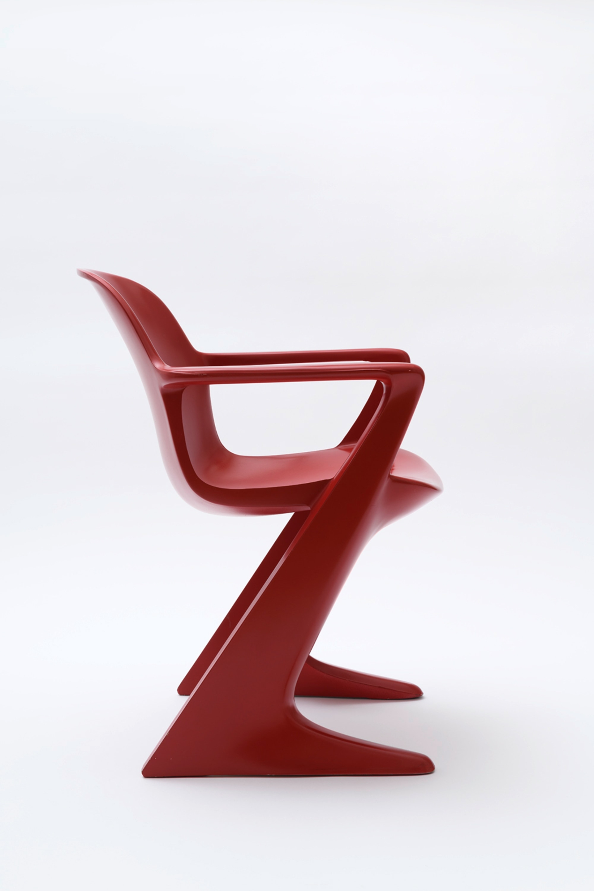 Ein roter Designerstuhl aus Kunststoff, fotografiert vor weißem Hintergrund. Der Stuhl hat eine markante Z-förmige Silhouette in der Seitenansicht. Die dynamische Form besteht aus einem durchgehenden, geschwungenen Stück, das von der Rückenlehne über die integrierten Armlehnen bis zum Standfuß reicht. Der Stuhl ist aus glänzendem, rotem Polyurethan gefertigt und zeigt eine futuristische, skulpturale Formensprache. Die minimalistische Gestaltung verbindet Funktionalität mit ästhetischer Innovation. Die Aufnahme präsentiert den Stuhl in einer klaren Studiofotografie, die die markante Form des Möbelstücks betont.