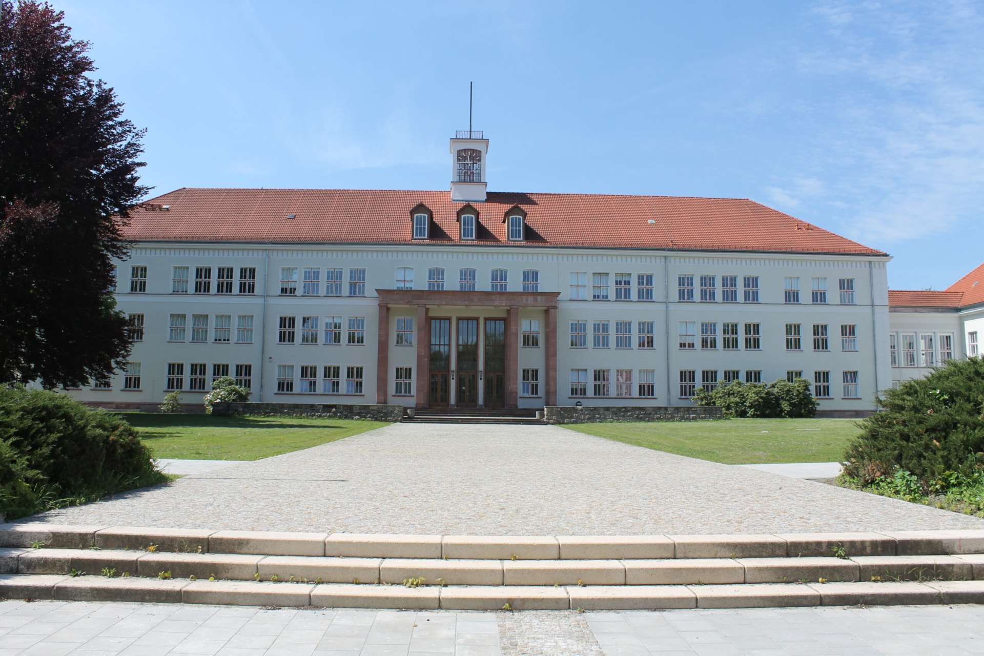 Frontalansicht der Gesamtschule 3 in Eisenh&uuml;ttenstadt - ein imposantes dreist&ouml;ckiges wei&szlig;es Geb&auml;ude im klassischen Schulbaustil mit rotem Ziegeldach, zentralem Eingangsportal und charakteristischem Uhrenturm, umgeben von gepflegten Gr&uuml;nanlagen und einem breiten Zugangspfad mit Treppenstufen