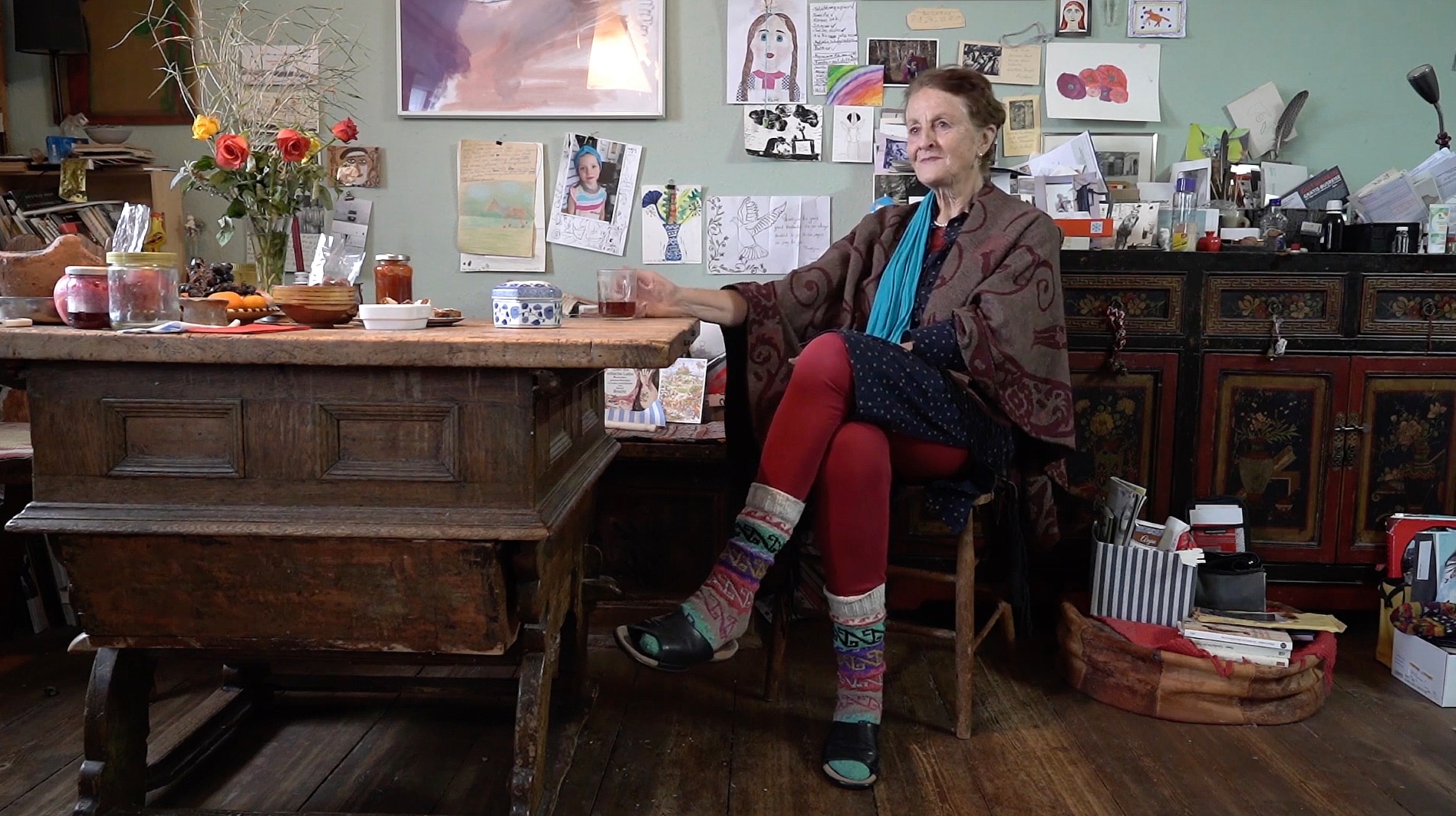Linde Bischof in ihrem gem&uuml;tlichen Wohnraum mit bunten Socken, roter Strumpfhose und einem Schal, umgeben von Kinderzeichnungen und einem antiken Holztisch