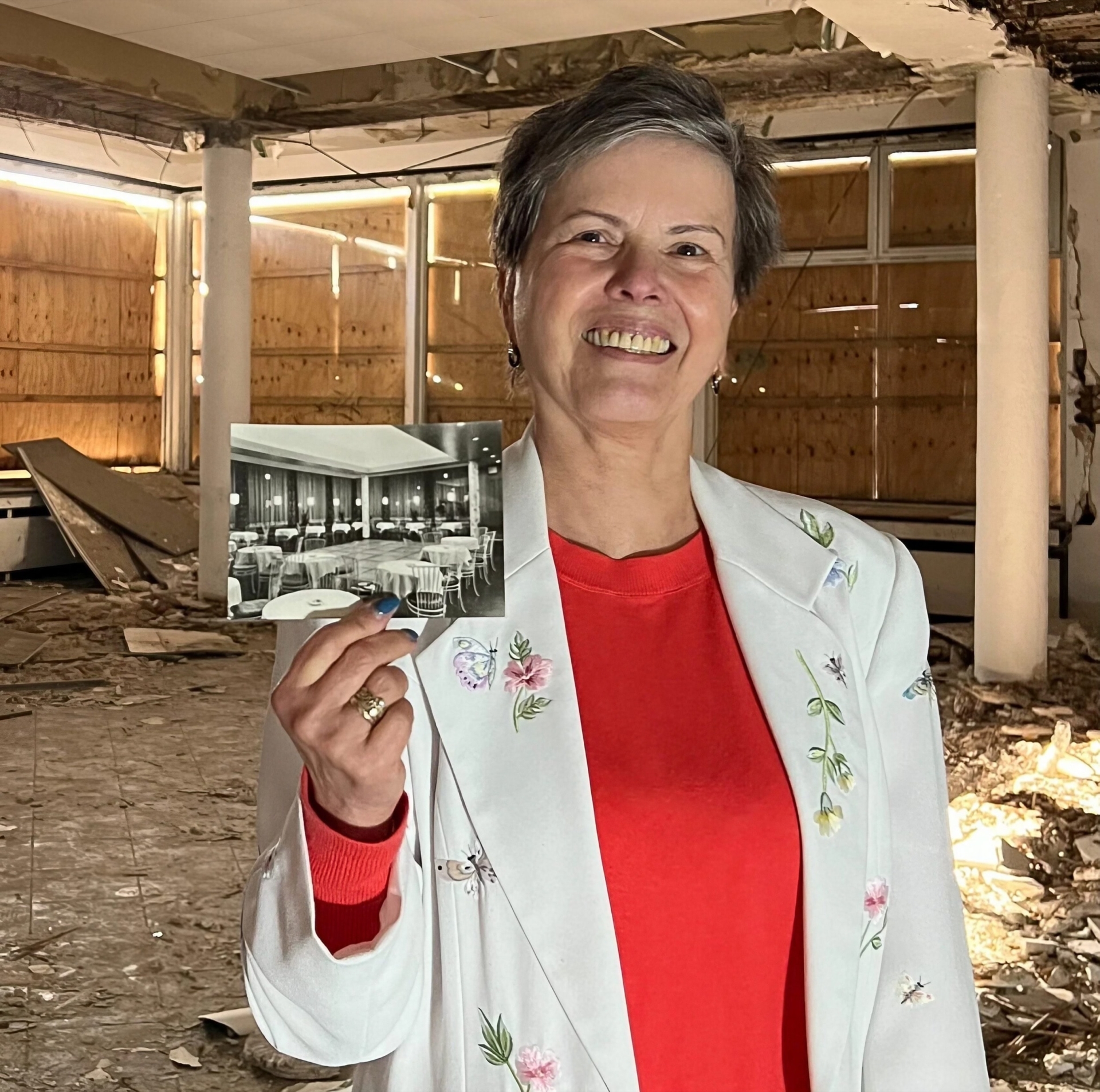 Frau Krause in rotem Shirt und wei&szlig;er Blumenjacke h&auml;lt ein historisches Foto in einem verfallenen Geb&auml;ude.