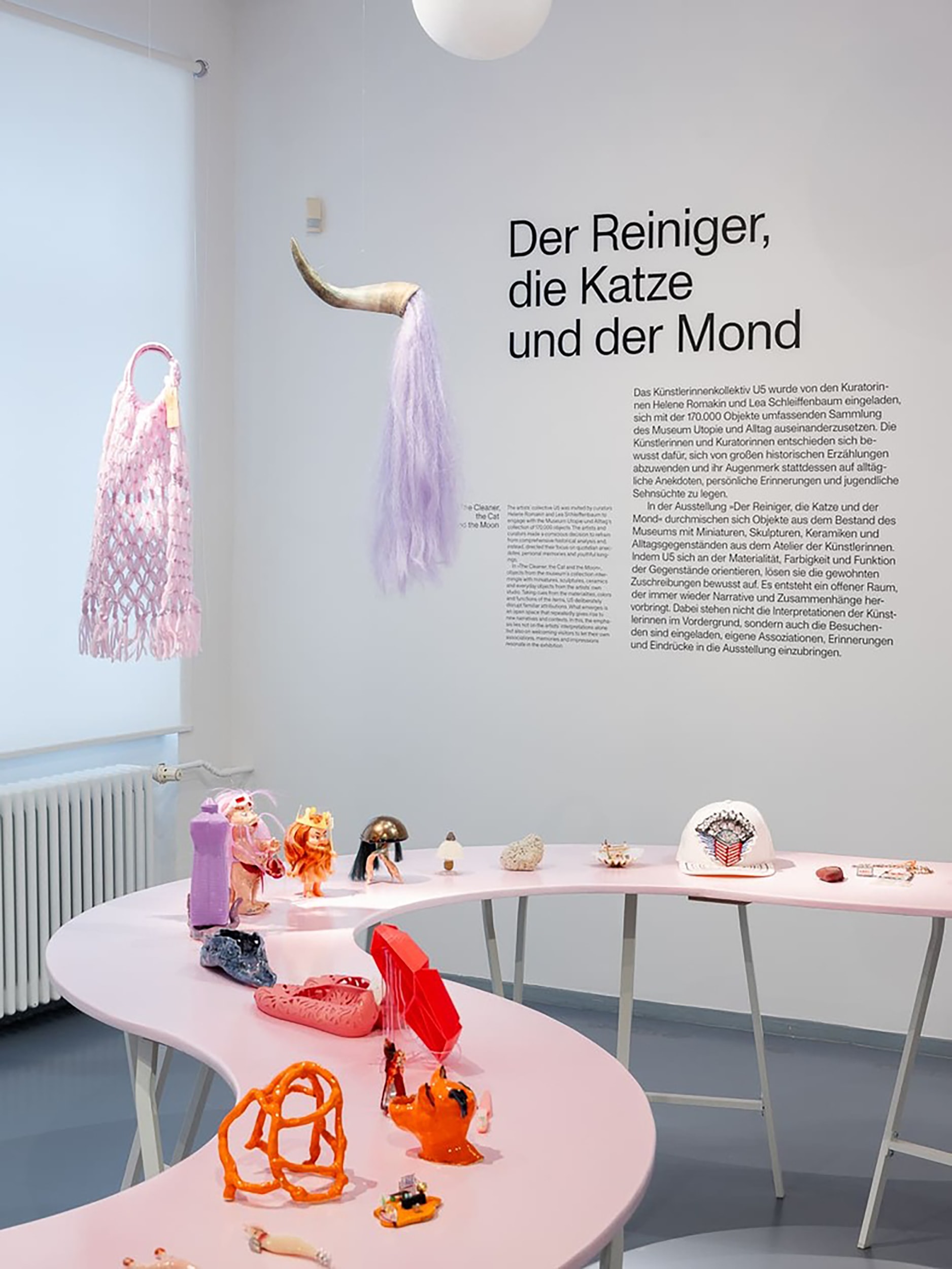 Ausstellungsansicht 'Der Reiniger, die Katze und der Mond' mit geschwungenem Tisch, auf dem kleine Skulpturen pr&auml;sentiert werden, an der Wand h&auml;ngen textile Kunstwerke
