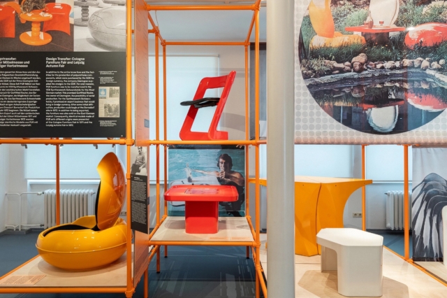 Im Vordergrund sind innovative Möbelstücke der 1970er Jahre zu sehen, präsentiert auf einem orangefarbenen Regalsystem. Links ein auffälliger gelb-oranger Sessel in organischer Form, in der Mitte ein roter Klappstuhl mit geometrischem Design, und rechts ein weißer Hocker. An den Wänden befinden sich Informationstafeln und historische Fotografien, die den Kontext der Designobjekte erläutern. Die Ausstellung ist in einem hellen Raum mit Heizkörpern an den Wänden installiert und zeigt die Entwicklung des DDR-Designs mit Fokus auf Polyurethan-Möbel. Im Hintergrund sind großformatige Ausstellungsbilder zu erkennen, die weitere Designobjekte dokumentieren.