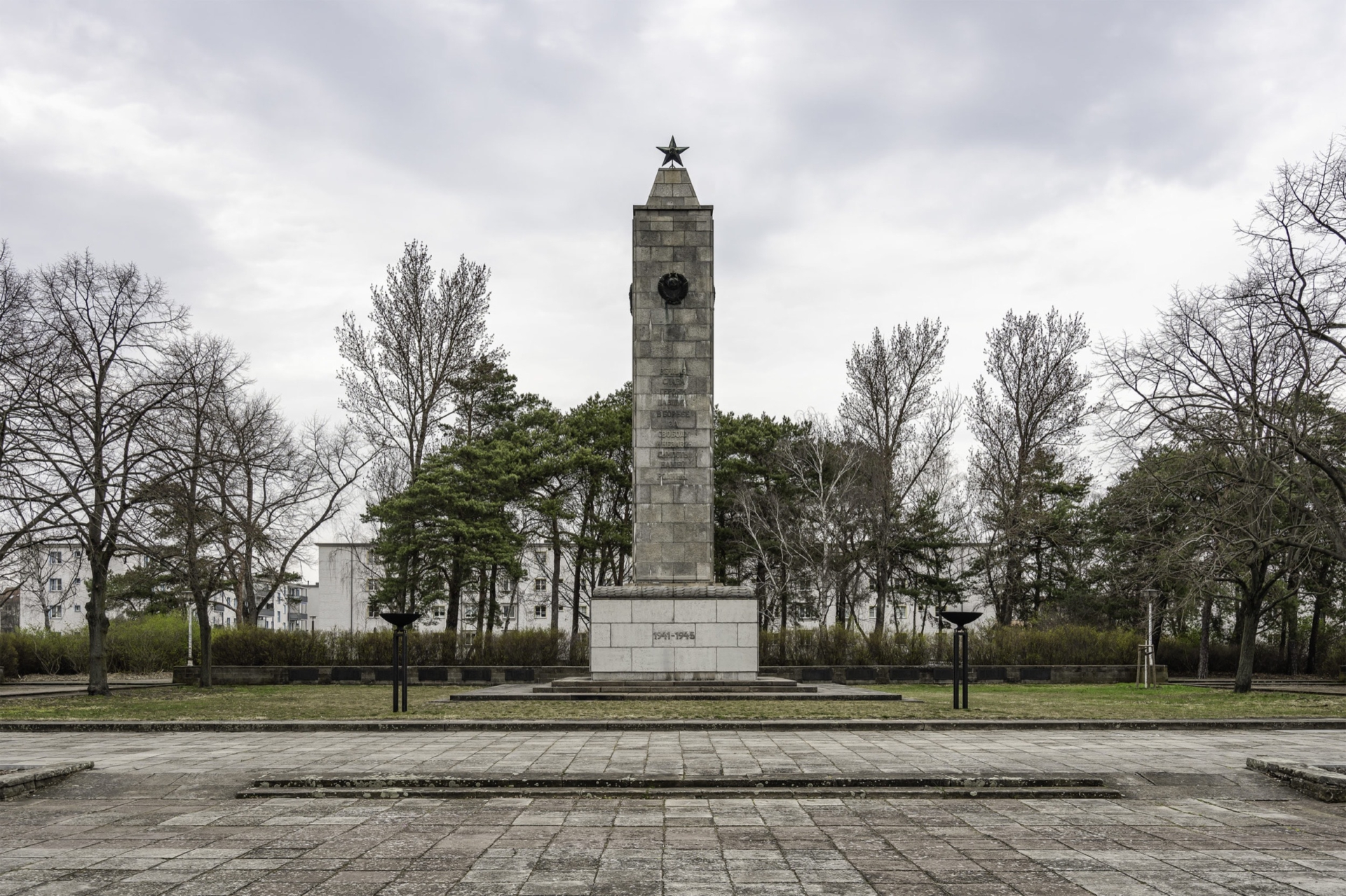 Zentraler Gedenkstein in Form eines hohen Obelisken aus hellem Stein, mit einem Sowjetstern an der Spitze und einem Medaillon in der Mitte. Der Obelisk steht auf einem gepflasterten Platz, umgeben von B&auml;umen und &auml;lteren Wohngeb&auml;uden im Hintergrund.