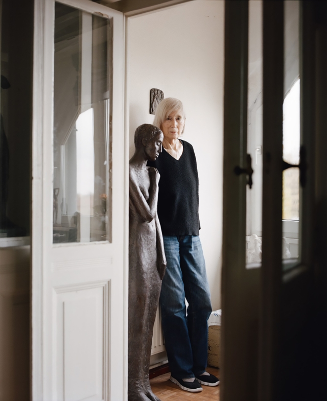 Carin Kreuzberg in schwarzem Pullover und Jeans steht neben einer lebensgroßen Bronzeskulptur einer schlanken Figur vor weißer Wand