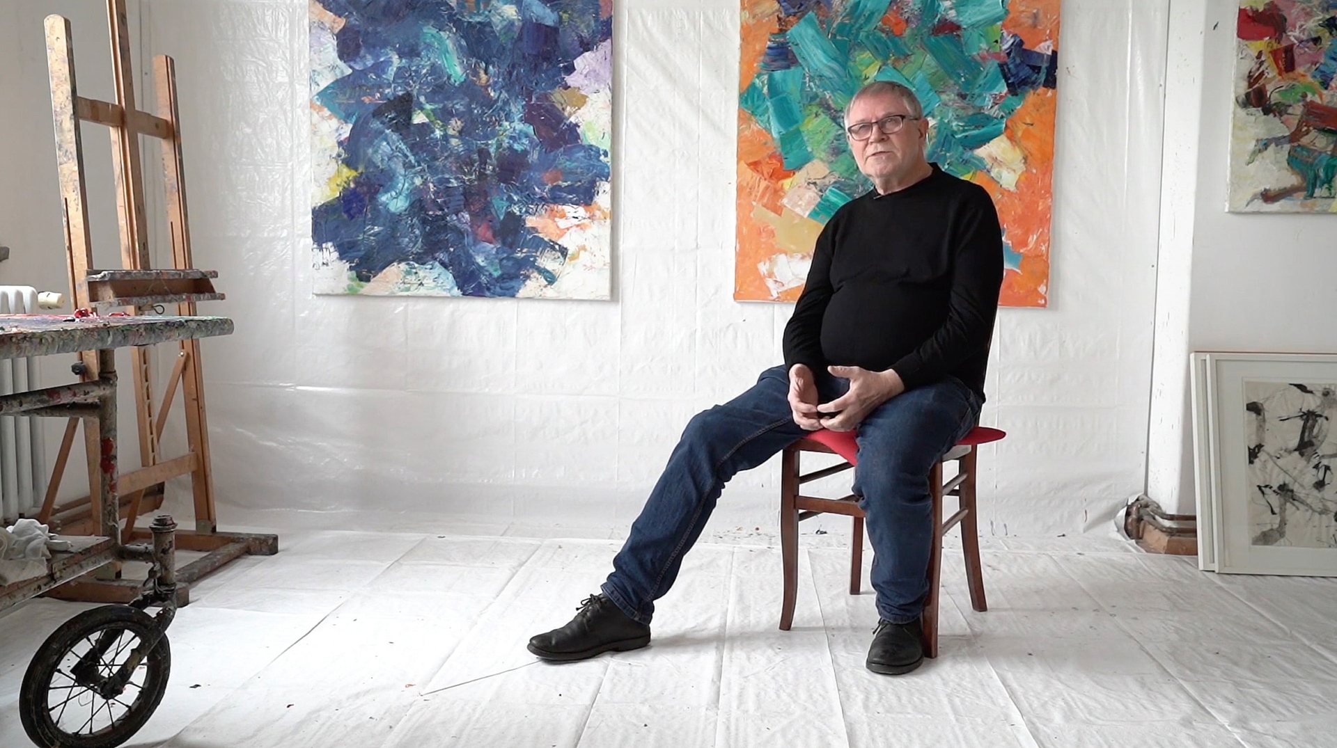 Volker Henze im schwarzen Pullover sitzt auf rotem Stuhl in einem K&uuml;nstleratelier mit abstrakten Gem&auml;lden an den W&auml;nden.