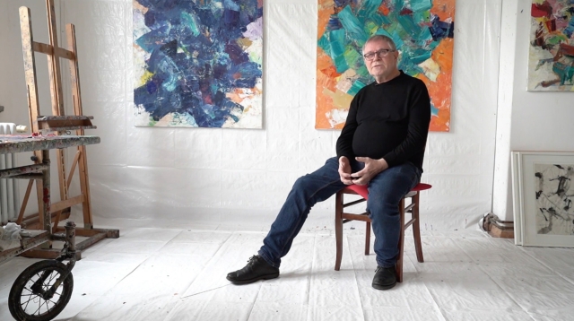 Volker Henze im schwarzen Pullover sitzt auf rotem Stuhl in einem Künstleratelier mit abstrakten Gemälden an den Wänden.
