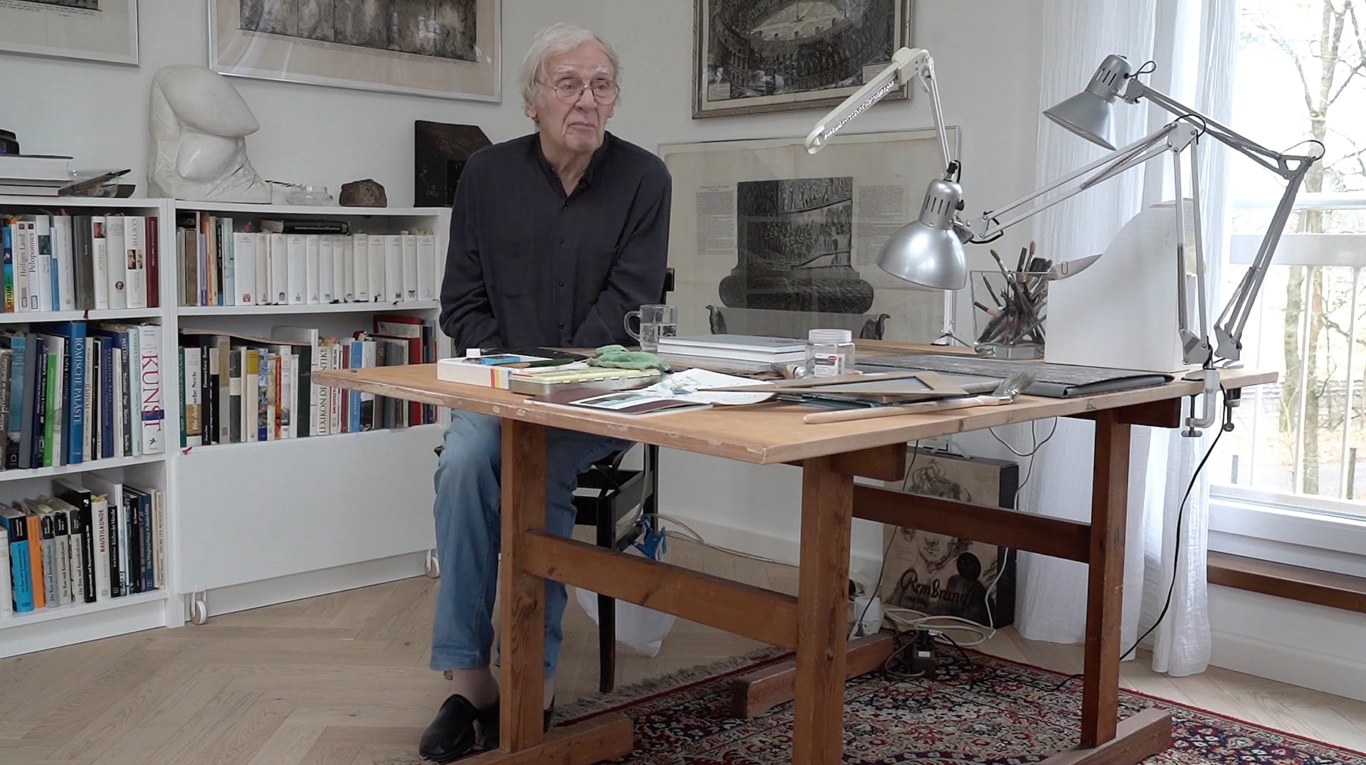 Walter Herzog an seinem Arbeitstisch mit Schreibtischlampen, umgeben von B&uuml;cherregalen und Kunstwerken an den W&auml;nden