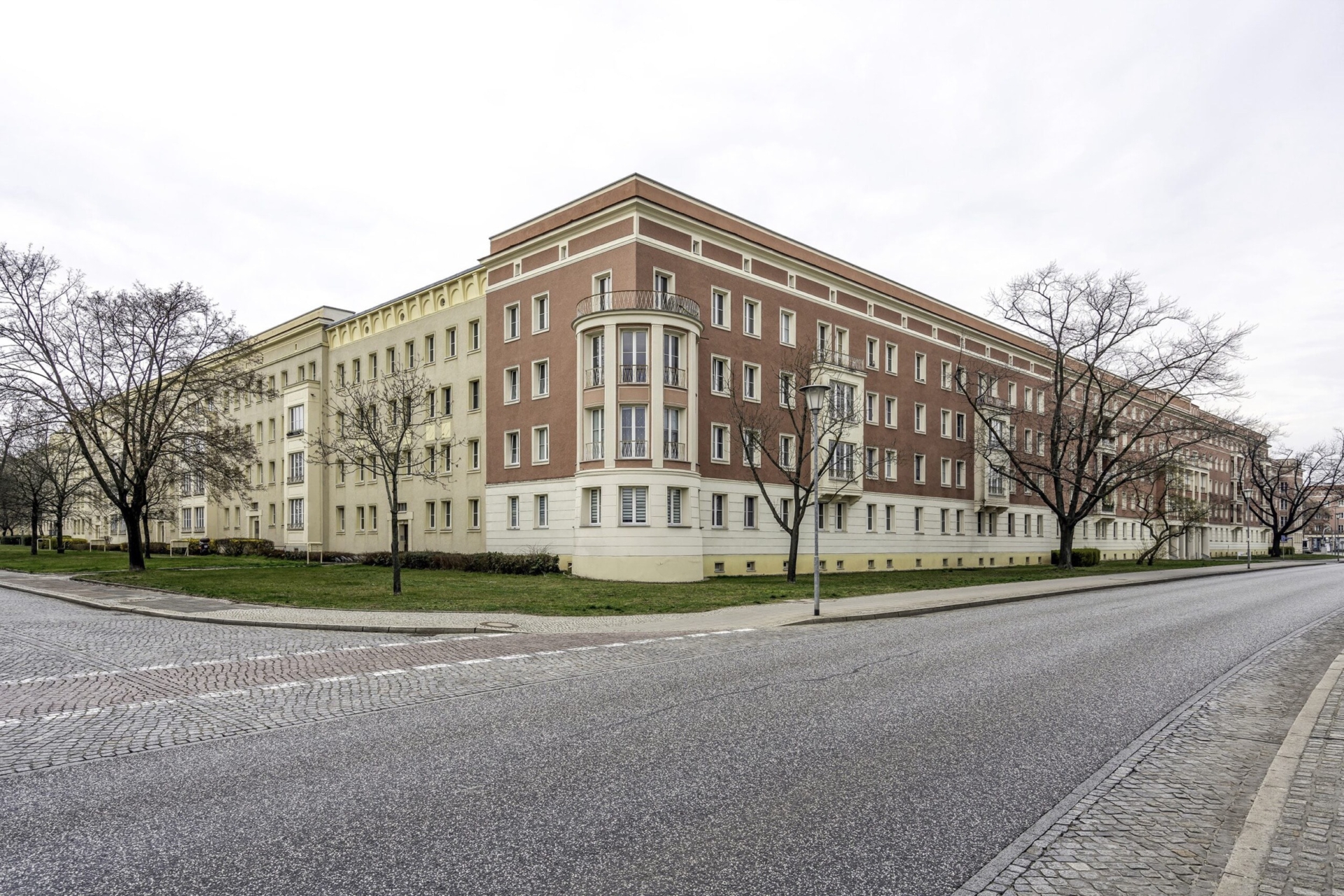 Mehrgeschossiger Wohnblock mit hellbeiger und roter Fassade, geschwungener Eckbebauung und symmetrisch angeordneten Fenstern. Das Geb&auml;ude steht an einer breiten Stra&szlig;e mit Kopfsteinpflaster-Randstreifen, flankiert von B&auml;umen und kleinen Gr&uuml;nfl&auml;chen.