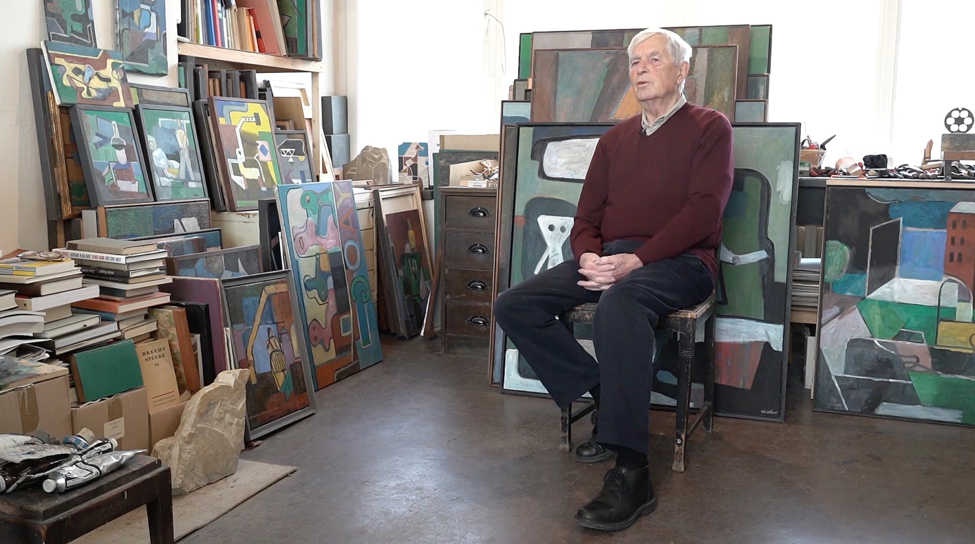 Wolfgang Leber sitzt in seinem Atelier auf einem Stuhl, umgeben von zahlreichen seiner geometrisch-abstrakten Gem&auml;lde und Kunstb&uuml;chern