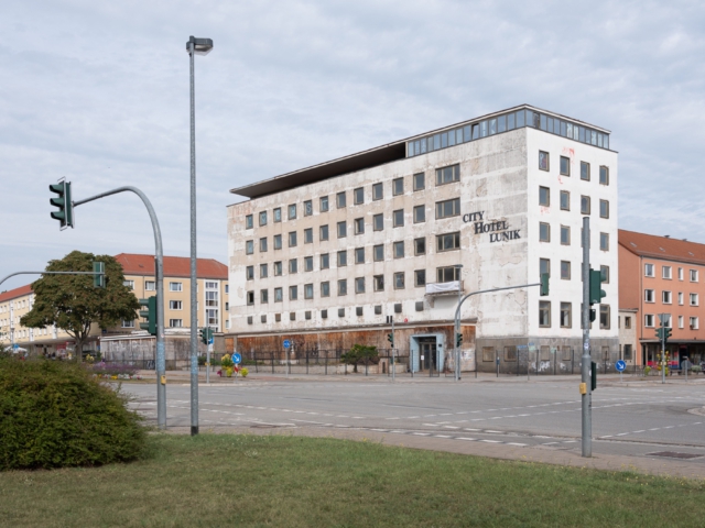 Sechsgeschossiges City-Hotel Lunik in Eisenh&uuml;ttenstadt, ein charakteristischer Hotelbau der DDR-Moderne aus den 1960er Jahren. Die wei&szlig;e Betonfassade zeigt eine streng geometrische Fensteranordnung und tr&auml;gt noch den originalen Schriftzug. Der funktionalistische Bau steht heute leer und weist deutliche Verfallsspuren auf, umgeben von typischer DDR-Wohnbebauung mit ockerfarbenen Fassaden.