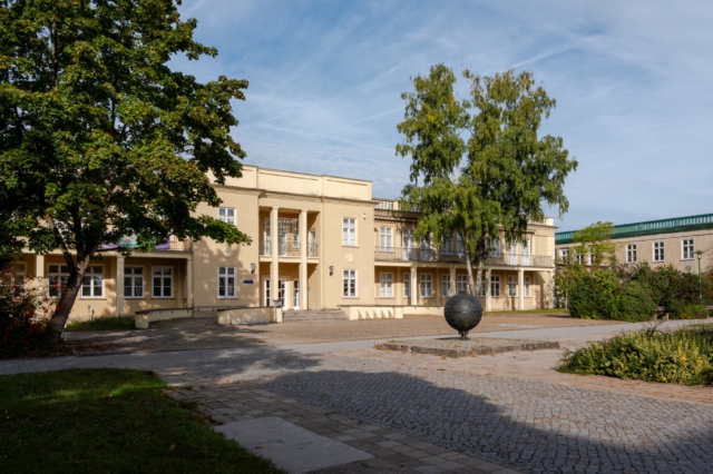 Aktuelle Aufnahme des ehemaligen DDR-KIndergrippe im Morgenlicht. Die cremefarbene Fassade mit klassizistischen Elementen erstreckt sich symmetrisch, mit zentralem S&auml;uleneingang und schmiedeeisernen Balkongel&auml;ndern. Auf dem gepflasterten Vorplatz steht eine schwarze Metallkugel als Kunstobjekt. Alte Birken und eine gepflegte Gr&uuml;nanlage rahmen das sanierte Geb&auml;ude ein, das die DDR-Architektur der 1950er Jahre repr&auml;sentiert.