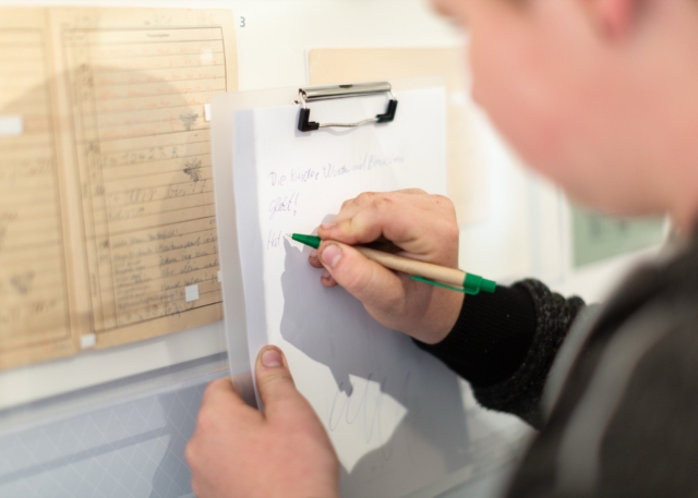 Eine Hand mit gr&uuml;nem Kugelschreiber schreibt auf ein wei&szlig;es Klemmbrett, im Hintergrund ist ein vergilbtes, liniertes Dokument aus DDR-Zeiten an der Wand zu erkennen. Die Aufnahme zeigt einen Moment der aktiven Auseinandersetzung mit historischen Dokumenten im Museumskontext.