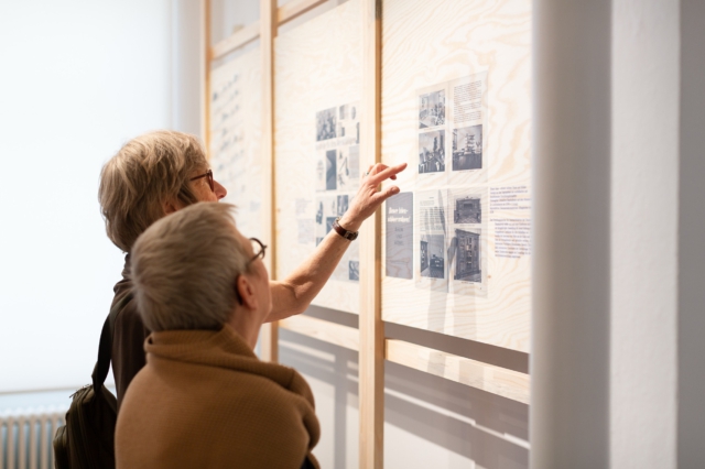Museumsbesucher:innen betrachten eine Ausstellungstafel mit historischen Schwarz-Wei&szlig;-Fotografien und Dokumenten zur DDR-Geschichte. Die in einem hellen Holzrahmen pr&auml;sentierte Dokumentation wird von warmem Tageslicht beleuchtet. Die sorgsam kuratierten Archivmaterialien zeigen typische Alltagsszenen und Stadtansichten aus der DDR-Zeit."
