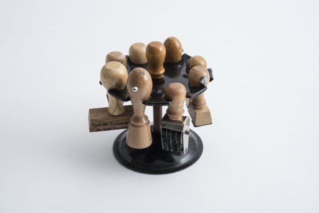 Rotierender Stempelhalter aus den 1970er Jahren. Der schwarze Kunststoffst&auml;nder tr&auml;gt mehrere Holzstempel in hellbrauner F&auml;rbung, kreisf&ouml;rmig angeordnet. Einer der sichtbaren Stempel tr&auml;gt die Aufschrift 'Stab der Zivilverteidigung', was die administrative Funktion des Objekts im DDR-Verwaltungsapparat dokumentiert.