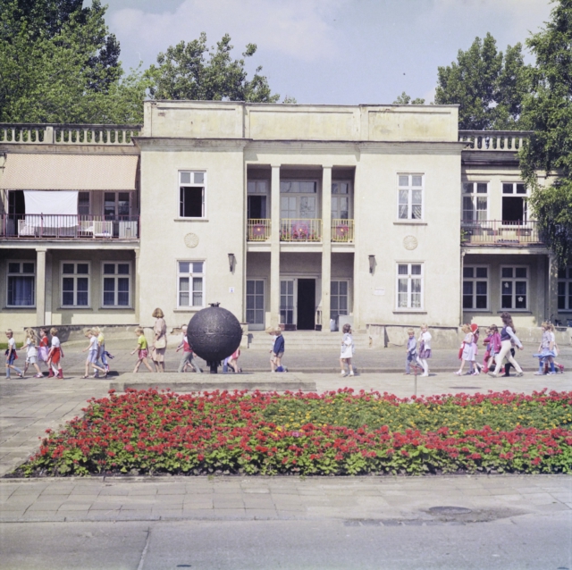Historische Aufnahme einer Kindergrippe. Eine Schulklasse in Sommerkleidung l&auml;uft am klassizistischen Geb&auml;ude vorbei. Im Vordergrund sind gepflegte Blumenbeete mit roten und gelben Bl&uuml;ten zu sehen, in der Mitte des Platzes steht eine schwarze Metallkugel als Kunstobjekt. Die cremefarbene Fassade mit S&auml;uleneingang und Balkonen zeigt die typische Architektur der 1950er Jahre.