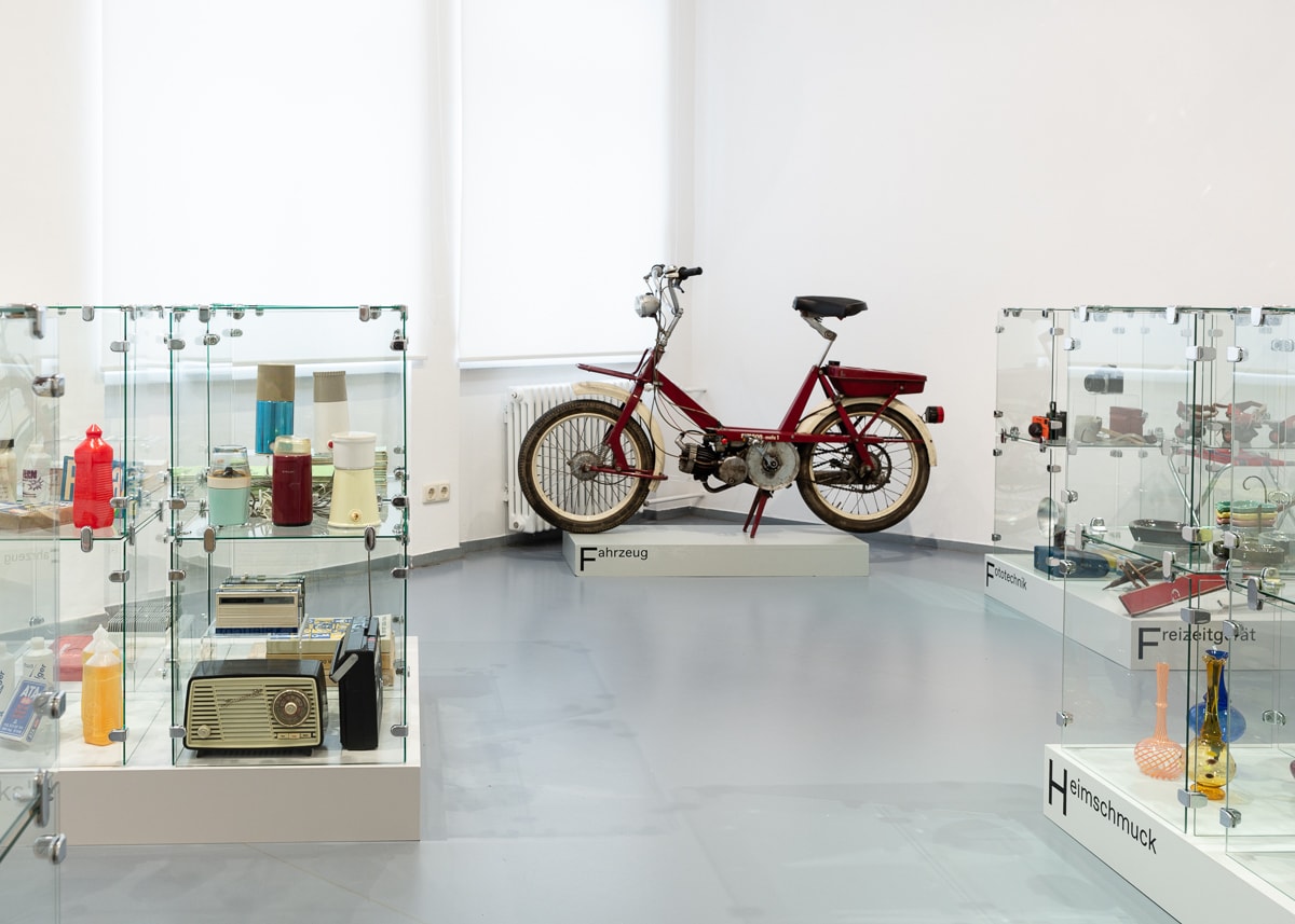Man blickt in einen Ausstellungsraum in Eisenh&uuml;ttenstadt. Links und rechts im Raum stehen Glasvitrinen. Viele verschiedene Objekte befinden sich darin. Hinten im Raum steht ein Simson Mofa.