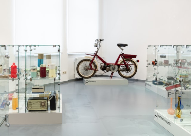 Man blickt in einen Ausstellungsraum in Eisenhüttenstadt. Links und rechts im Raum stehen Glasvitrinen. Viele verschiedene Objekte befinden sich darin. Hinten im Raum steht ein Simson Mofa.