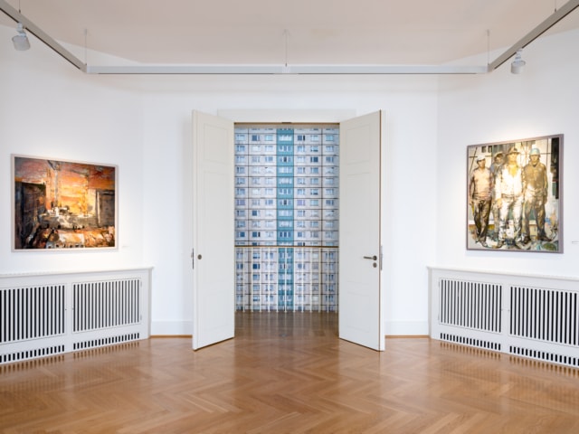 Man sieht einen Innenraum, der als Ausstellungsraum im Schloss Biesdorf genutzt wird. Der Boden besteht aus dunklem Holzparkett und die Wände sind weiß. An den Wänden hängt jeweils links und rechts ein Gemälde. Zwischen den Gemälden steht eine doppelflügelige Tür offen und gibt den Blick frei auf eine Fotografie die einen Plattenbau zeigt.