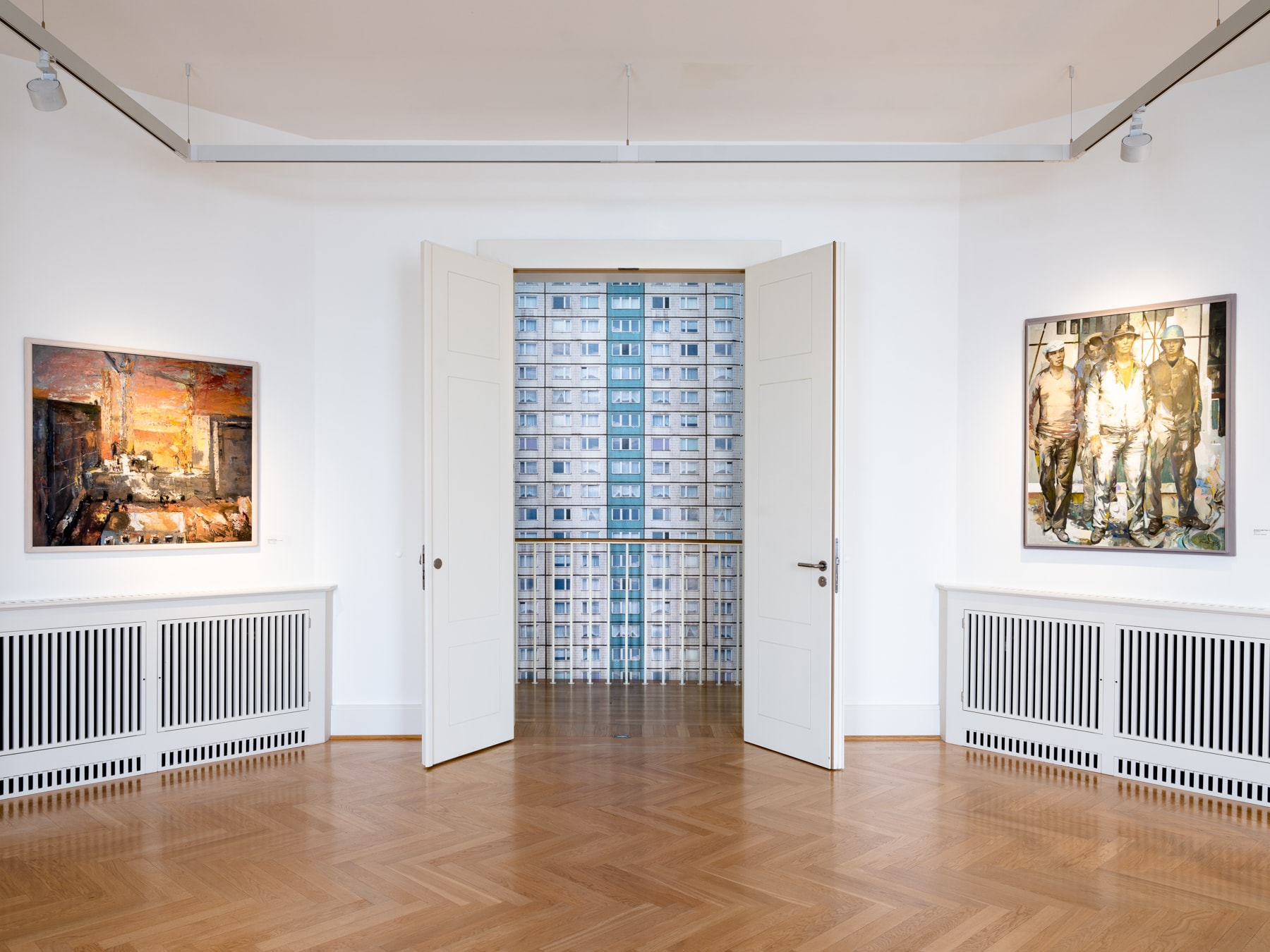 Man sieht einen Innenraum, der als Ausstellungsraum im Schloss Biesdorf genutzt wird. Der Boden besteht aus dunklem Holzparkett und die W&auml;nde sind wei&szlig;. An den W&auml;nden h&auml;ngt jeweils links und rechts ein Gem&auml;lde. Zwischen den Gem&auml;lden steht eine doppelfl&uuml;gelige T&uuml;r offen und gibt den Blick frei auf eine Fotografie die einen Plattenbau zeigt.
