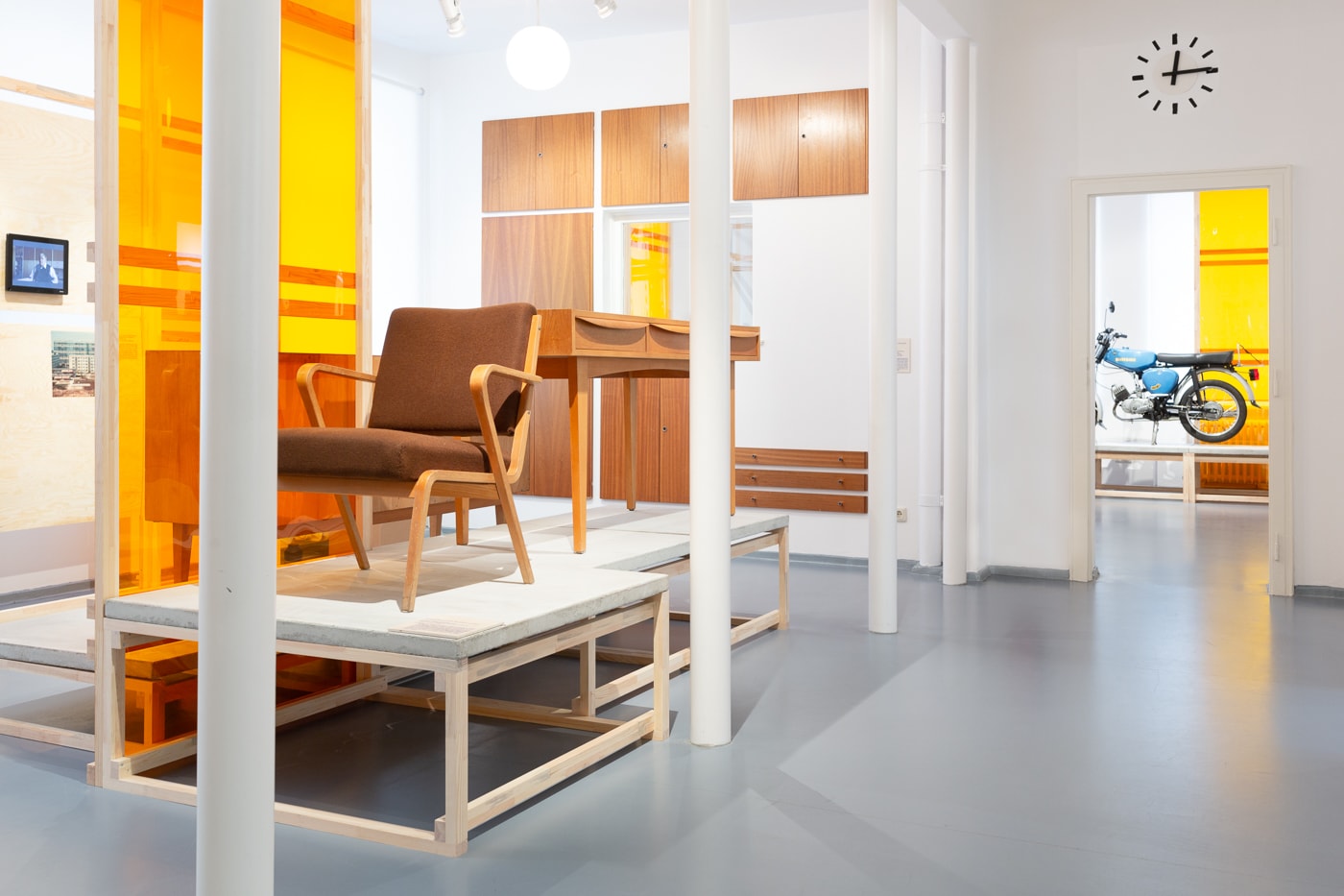 Zu sehen ist ein Ausstellungsraum in Eisenh&uuml;ttenstadt. Links sieht man Ausstellungsobjekte. Einen Sessel und dahinter einen Schreibtisch. Links daneben steht ein orangefarbener Raumtrenner Am rechten Bildrand l&auml;uft der Flur in den n&auml;chsten Ausstellungsraum entlang. Im Hintergrund des n&auml;chsten Raumes sieht man eine Simson S50 stehen.