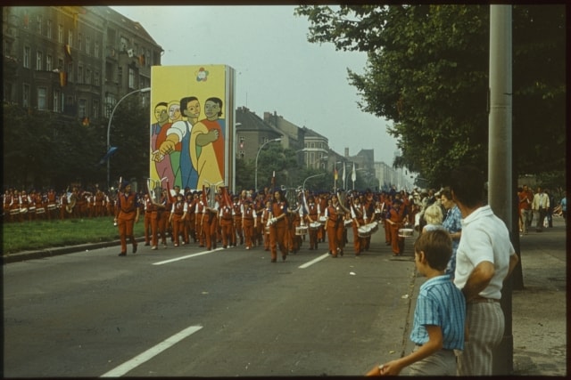 Eine farbige Fotografie. zusehen ist eine Parade mit Musikerinnen und Musiker in roten Uniformen. Am Straßenrand stehen Passanten. Auf einem Grünstreifen in der Mitte des Straßenzuges ist ein großes Plakat aufgestellt.