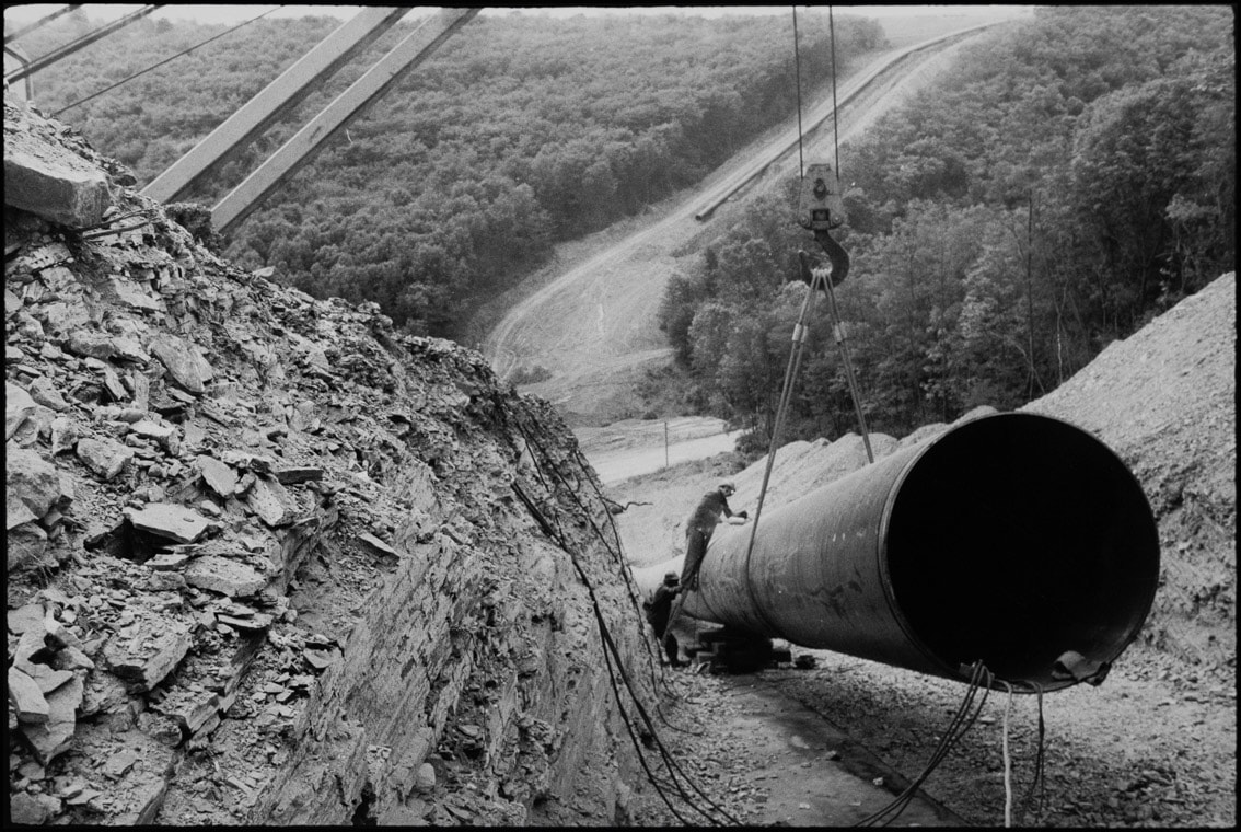 Auf dem schwarz-wei&szlig; Foto f&auml;llt der Blick von einem Berg aus hinunter in das Tal. Dabei ist der Berg zum Teil ausgeschachtet. Ein Kran und Bauarbeiter sind dabei ein Erdgasrohr in den Schacht einzubauen.