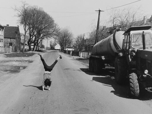 Schwarzweißfotografie. Landstraße auf der ein Kind einen Handstand macht. Am rechten Bildrand ist eine Güllewagen zu sehen.