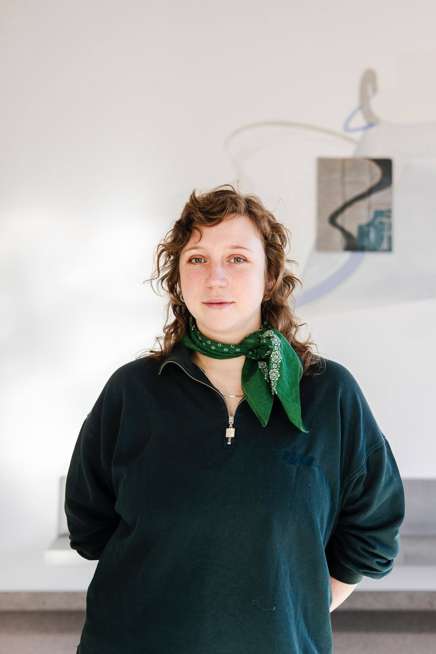 Eine Frau steht vor einem weißen Hintergrund. Sie trägt einen dunkelgrünen Pullover und trägt ein grünes Halstuch. Sie hat gelockte, schulterlange rotbraune Haare.